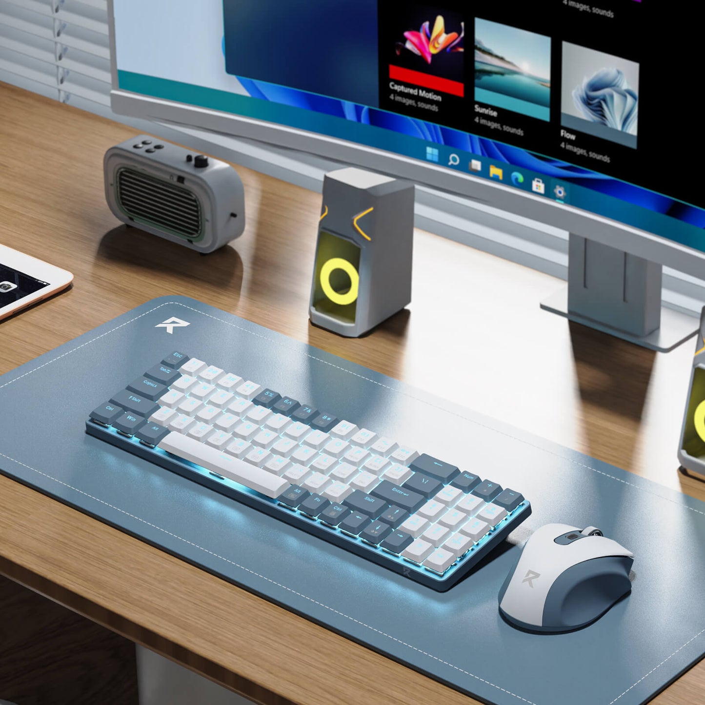 The keyboard and mouse are shown in a clean, modern office setup, with a monitor, speakers, and a portable speaker in the background.