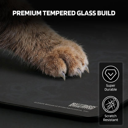 A cat's paw with claws tests the mouse pad's 'PREMIUM TEMPERED GLASS BUILD', marked as 'Super Durable' and 'Scratch Resistant' with Blizzard Entertainment branding.