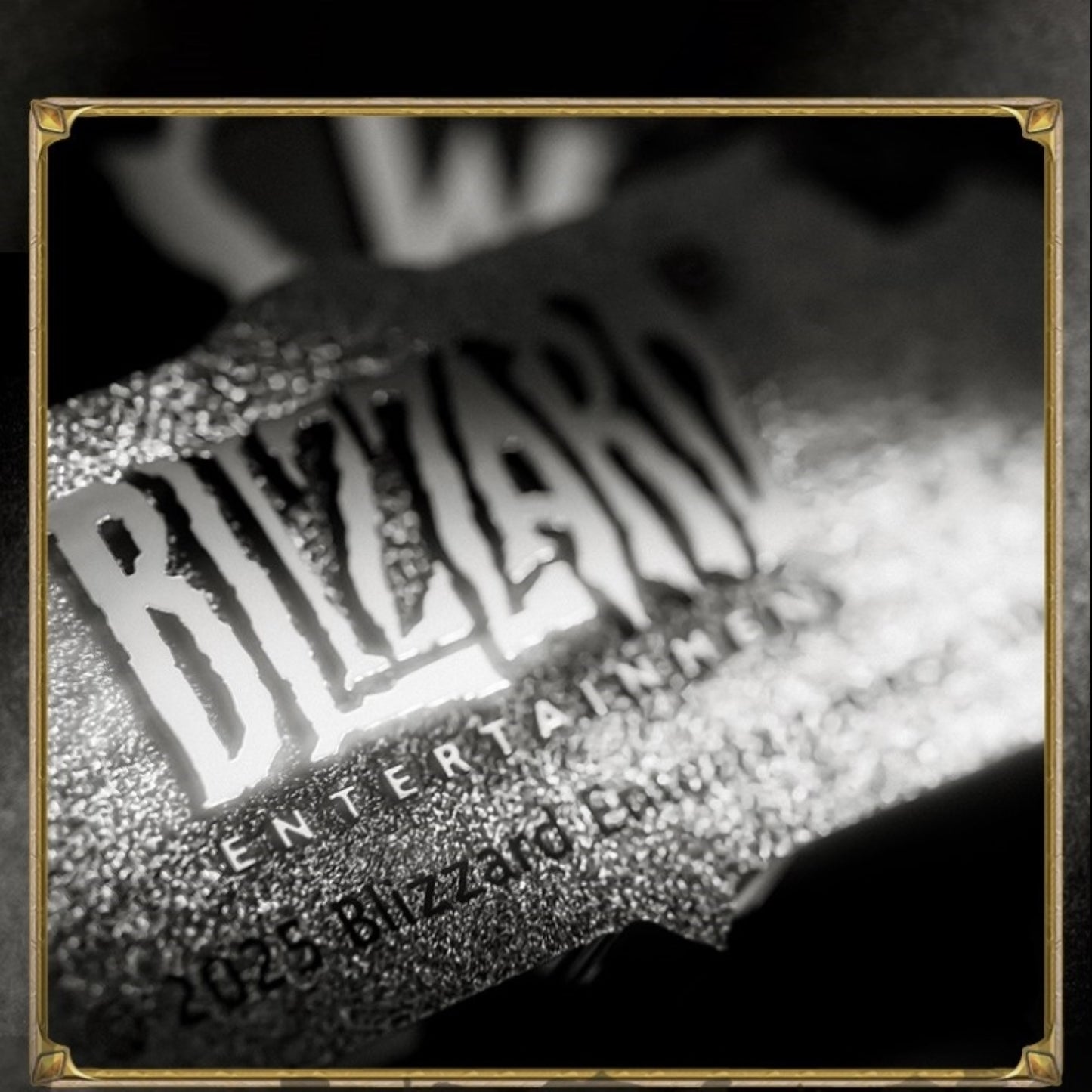 Within a gold-cornered frame, this black-and-white close-up shows a textured metallic emblem: bold "BLIZZARD" lettering glows under light, its grainy surface catching bright highlights against a softly blurred dark background.