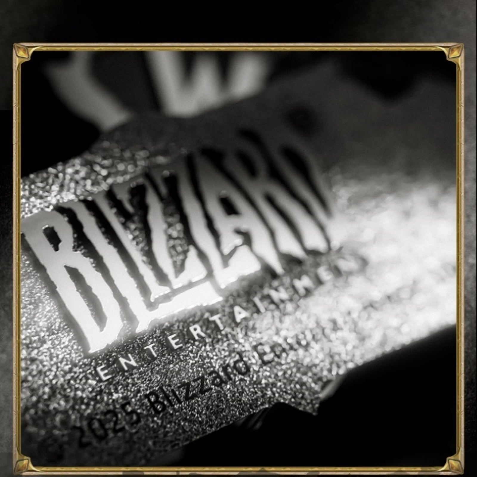 Within a gold-cornered frame, this black-and-white close-up shows a textured metallic emblem: bold "BLIZZARD" lettering glows under light, its grainy surface catching bright highlights against a softly blurred dark background.
