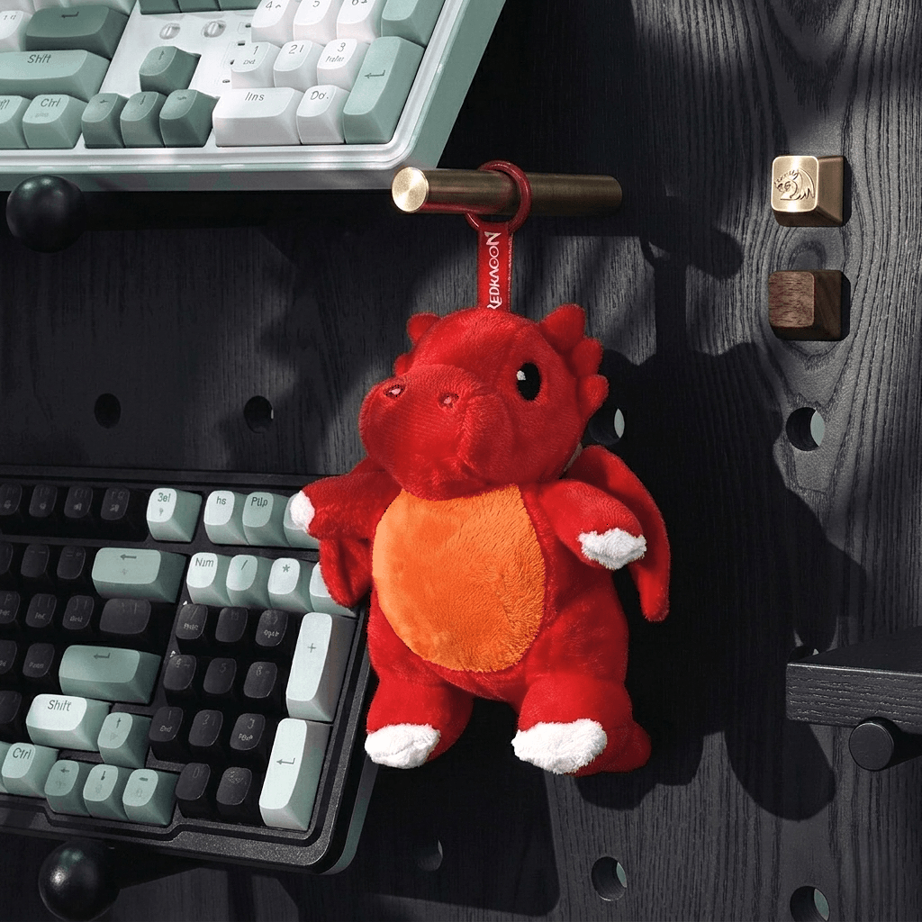 Red plush dragon toy hanging from a bag handle next to a keyboard on a wooden surface