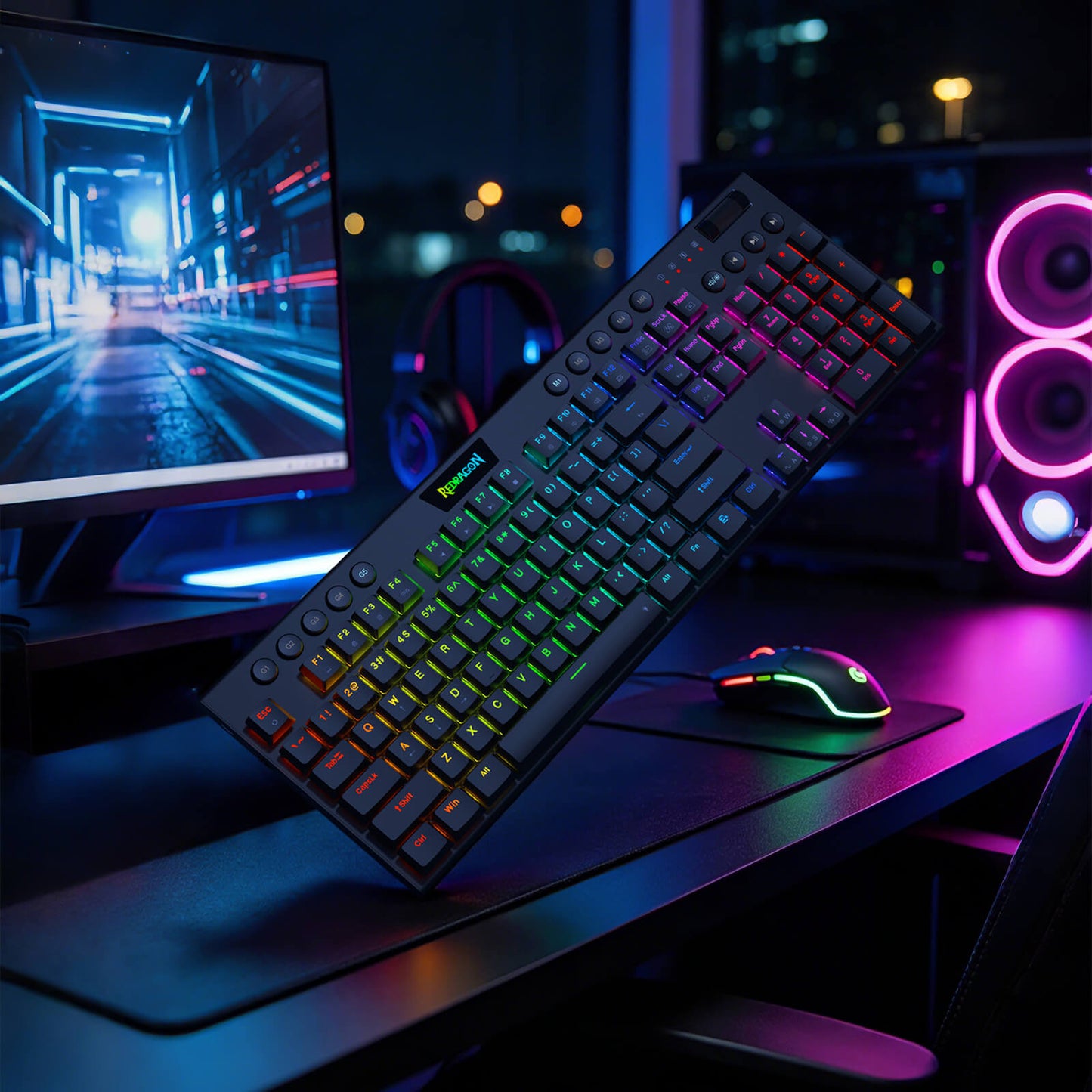 A black mechanical gaming keyboard with RGB backlighting is shown at an angle on a desk, with a matching RGB mouse and a computer monitor in a dark, modern gaming setup.