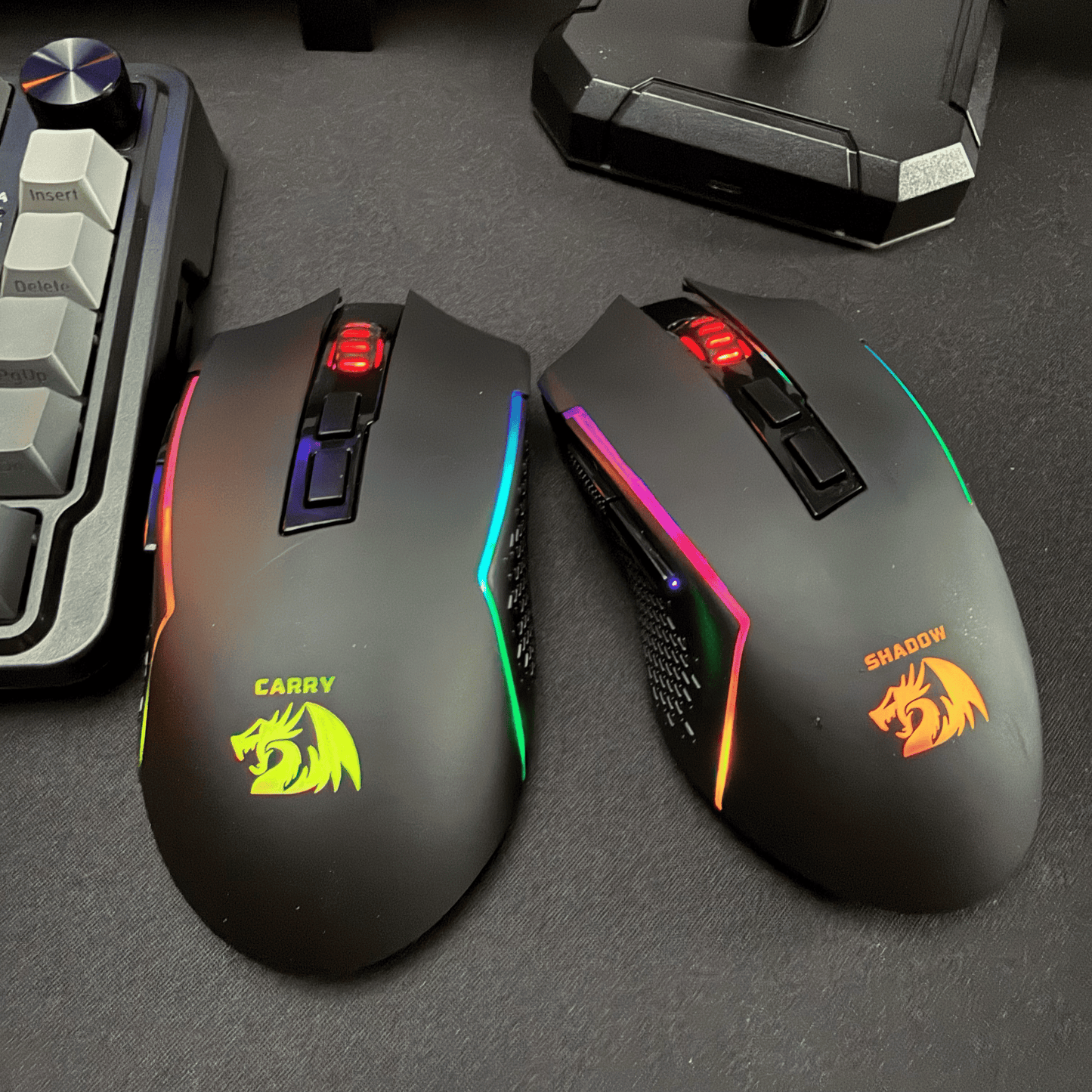 Two gaming mice with colorful lighting on a dark surface, surrounded by gaming accessories.