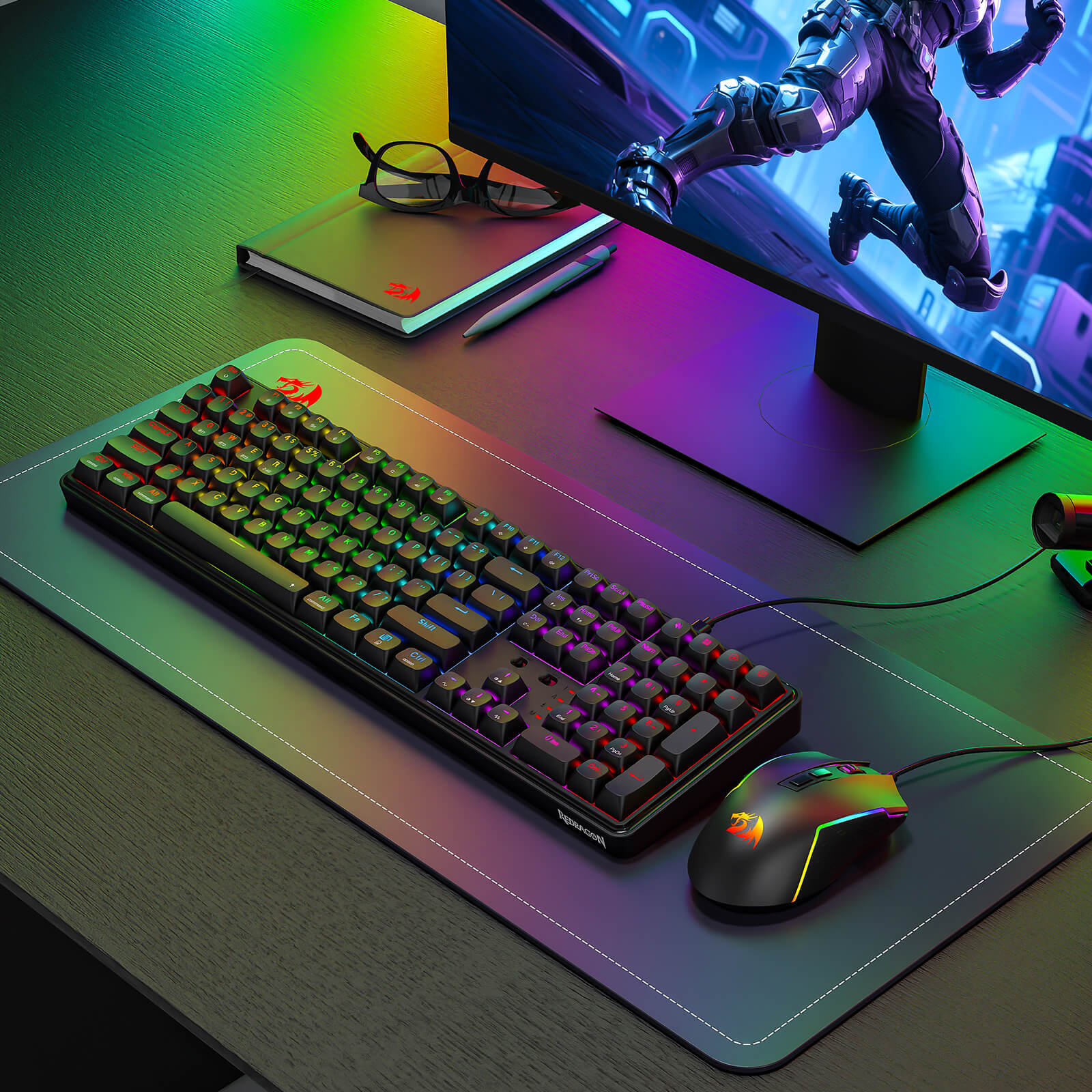 A Redragon RGB mechanical keyboard and gaming mouse are displayed on a branded desk mat in a modern gaming setup, with a monitor and webcam in the background.