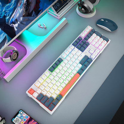 A white mechanical keyboard with multicolor RGB backlighting and custom keycaps, shown on a gray desk alongside a mouse, headphones, and other tech accessories.