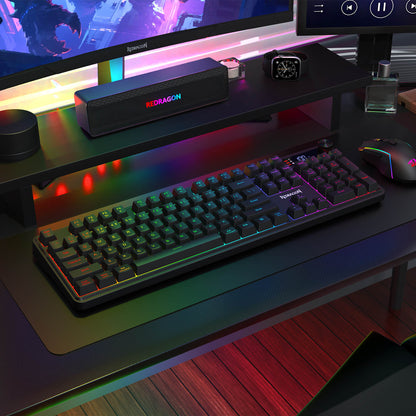 The Redragon RM104 Pro keyboard and RM104 mouse are displayed in a vibrant RGB-lit gaming setup, with a monitor, speakers, and a small potted plant.