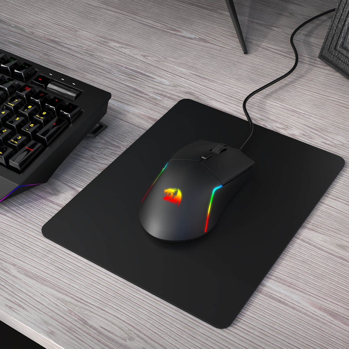 A Redragon RGB gaming mouse with a glowing logo and side strips rests on a black mouse pad next to a mechanical keyboard.