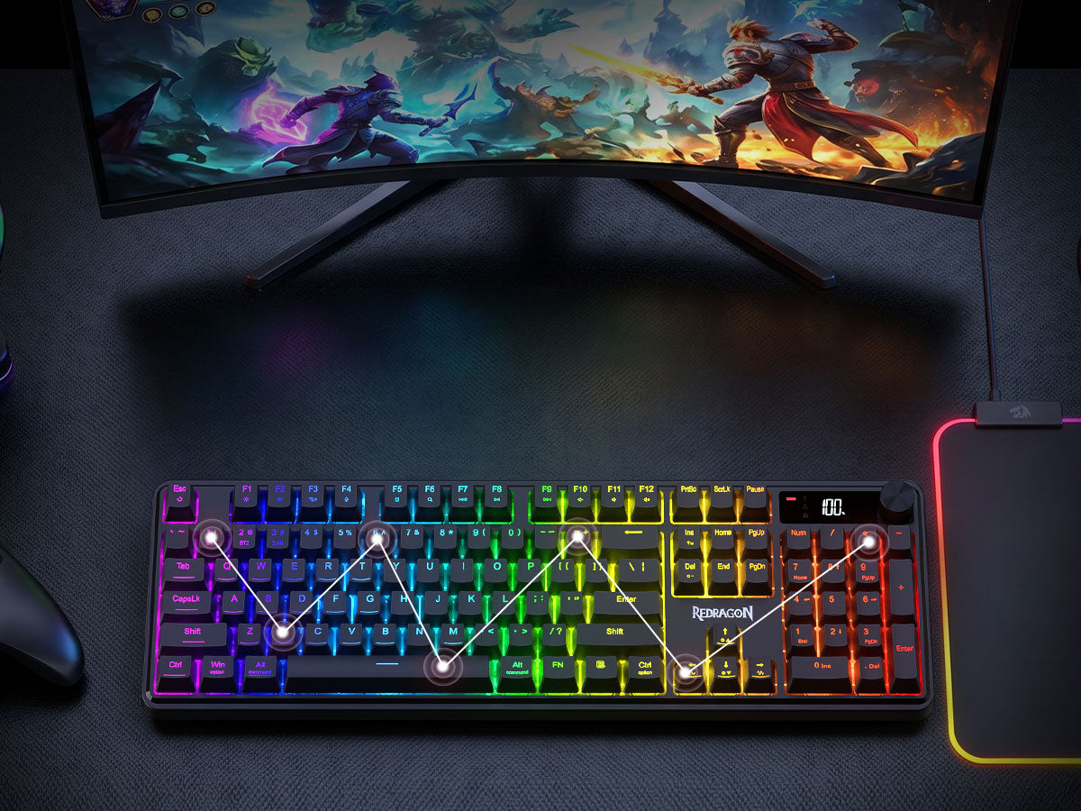 A compact mechanical gaming keyboard with vibrant RGB rainbow backlighting, shown on a dark desk in front of a curved monitor and next to a mouse pad.